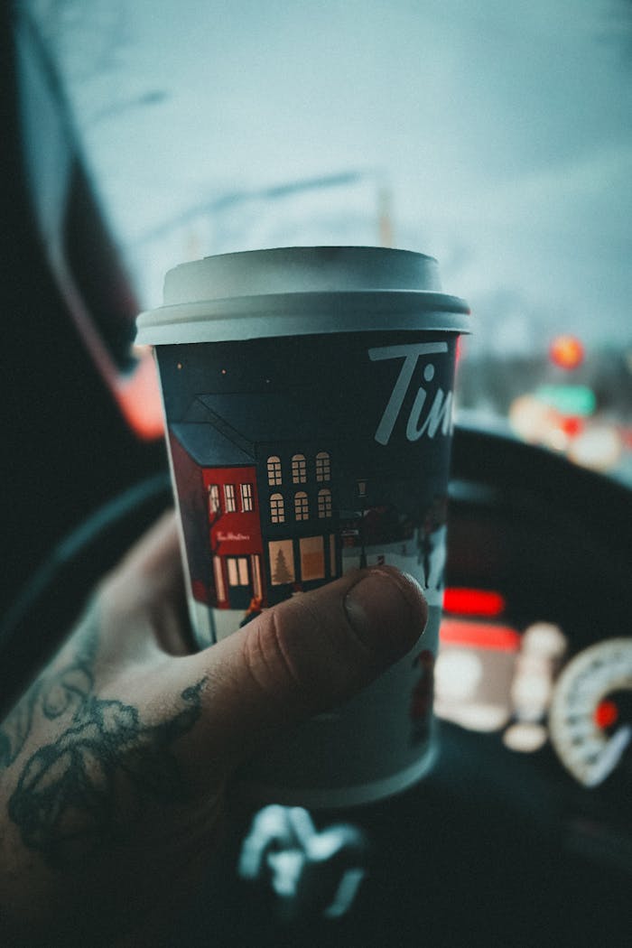 A tattooed hand holds a festive coffee cup inside a car, showcasing a winter atmosphere.